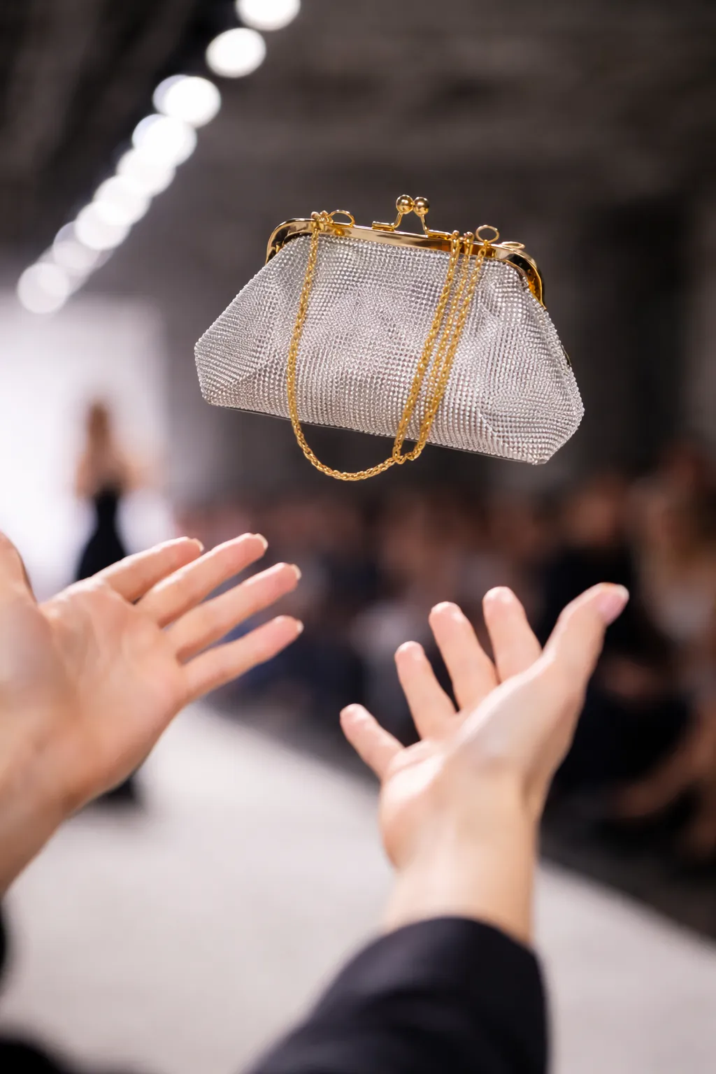 Silver crystal evening clutch with gold chain suspended above outstretched hands at a fashion runway show, capturing a luxury party lifestyle moment.